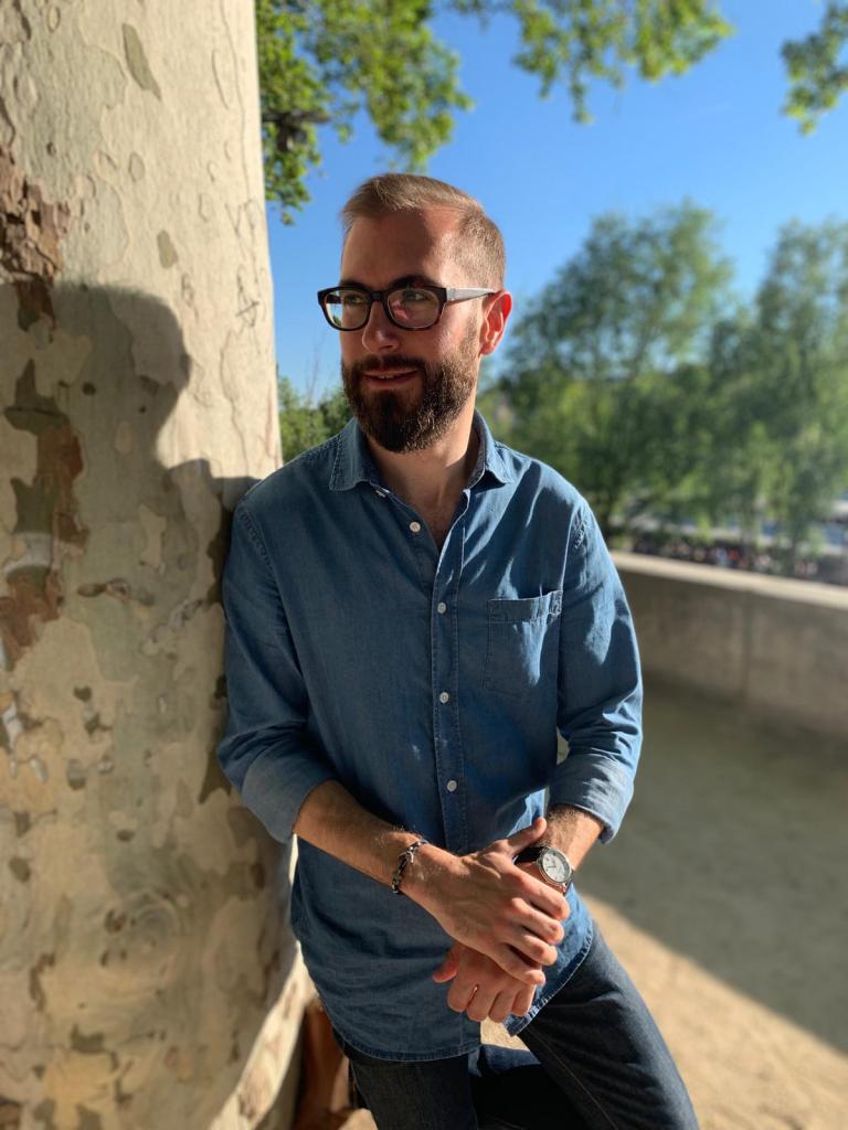 Joven con gafas y camisa de jean azul se apoya sobre un árbol mientras sostiene sus manos con reloj en la muñeca
