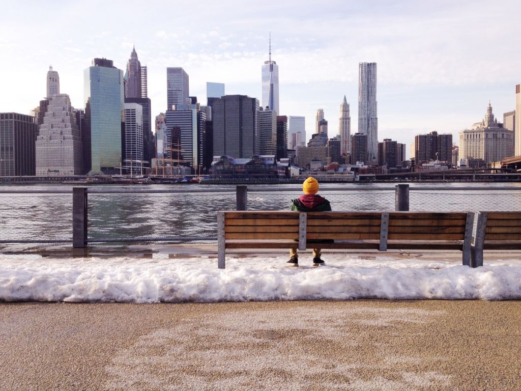 Personne assise sur un banc en hiver devant une rivière et les grattes ciel de New York